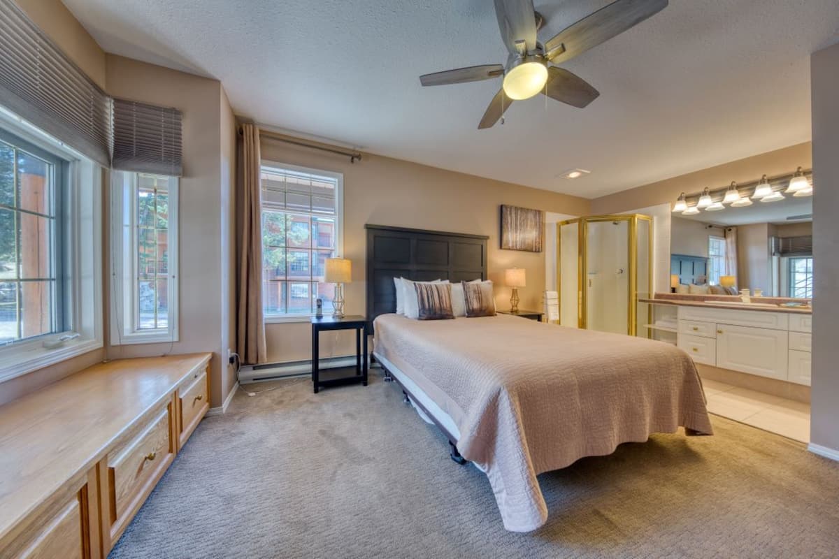 A bedroom with a large bed, a ceiling fan, a nightstand with lamps, a dresser, and a bathroom with a mirror and lights.