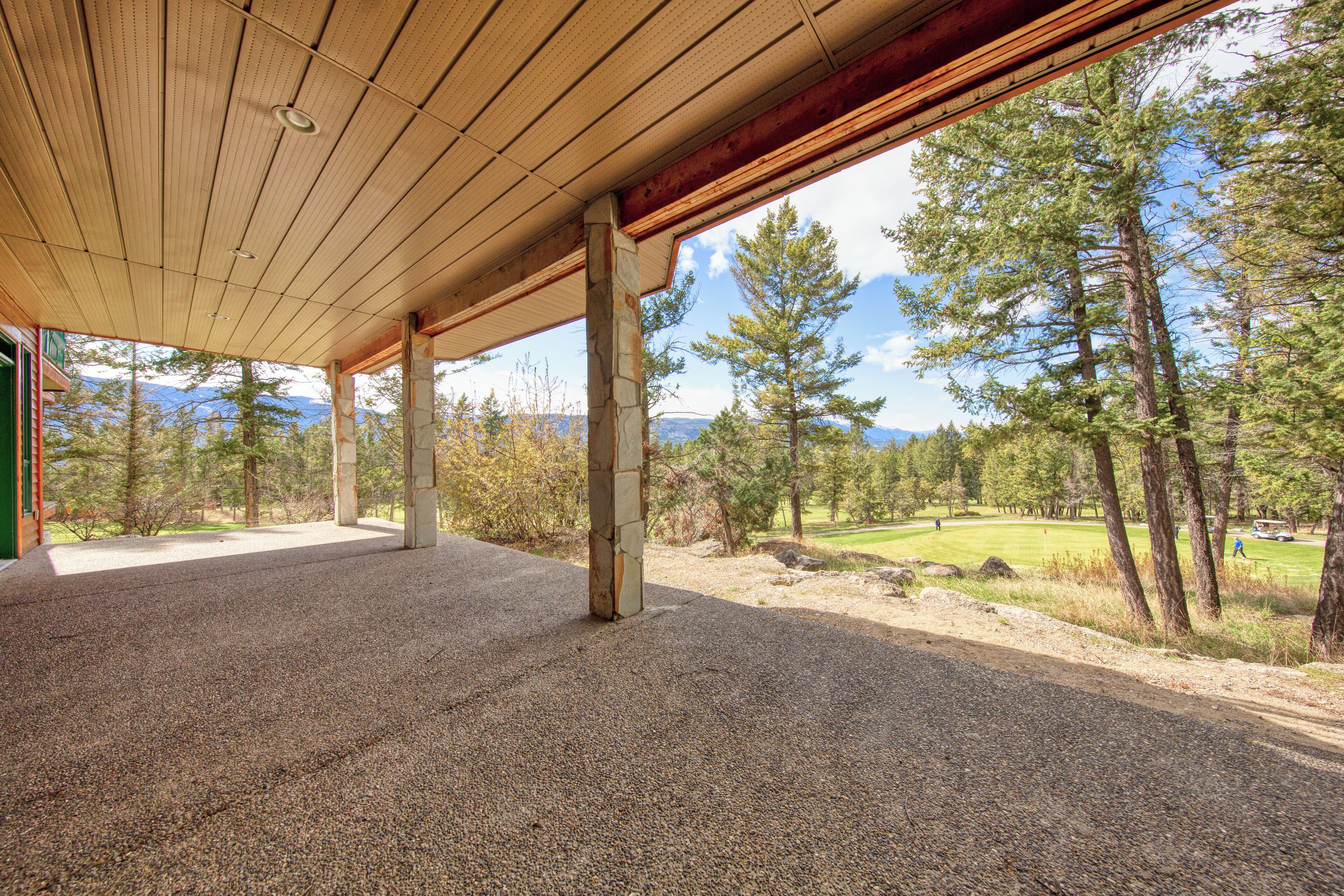 A covered outdoor area with a gravel floor, wooden beams, and a view of a golf course and surrounding trees.