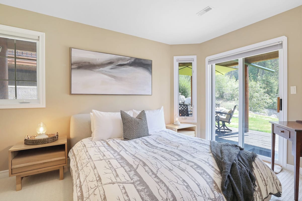 A bedroom with a bed, a bedside table with a lamp, a large window, and a view of the outdoors.