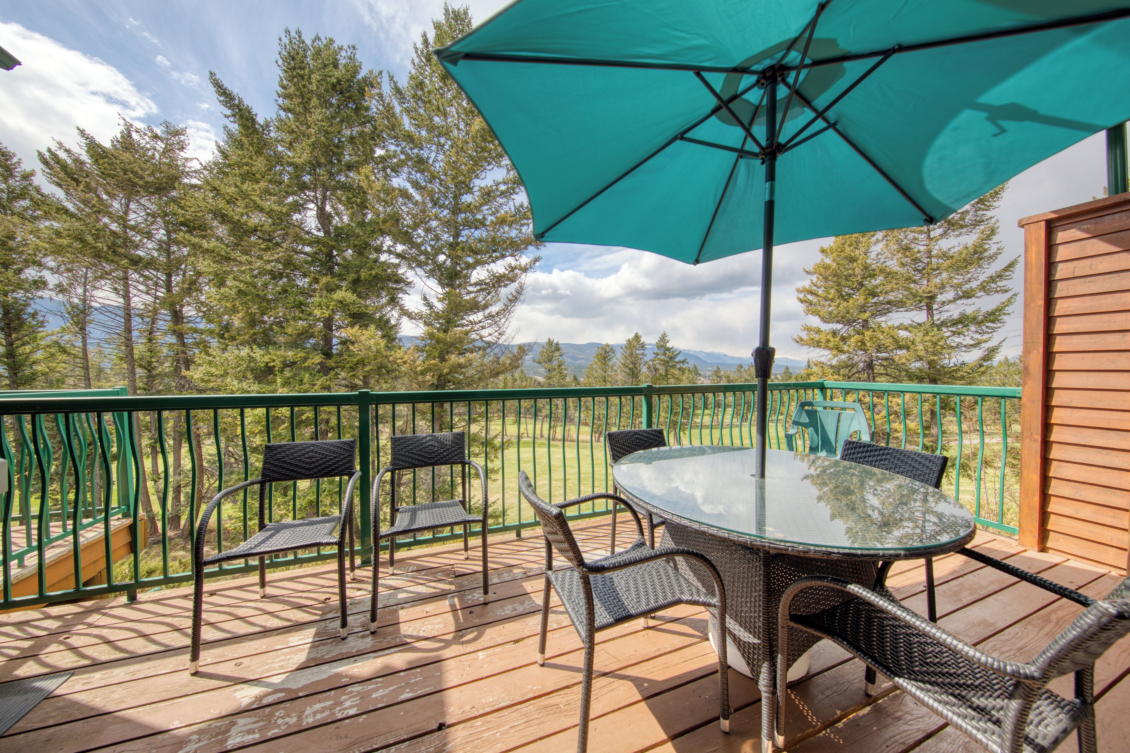 Une terrasse en bois avec une table et des chaises en verre, entourée d’une rambarde verte, donnant sur une zone boisée.