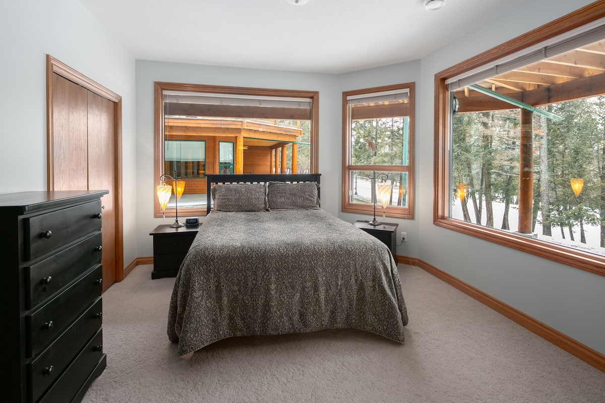 A bedroom with a large bed, wooden headboard, two nightstands, a dresser, and large windows offering a view of a wooden deck and trees.