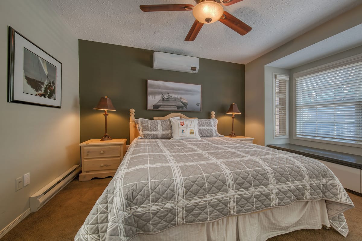 A bedroom with a bed, nightstands, a ceiling fan, wall-mounted air conditioning, and a window with blinds.