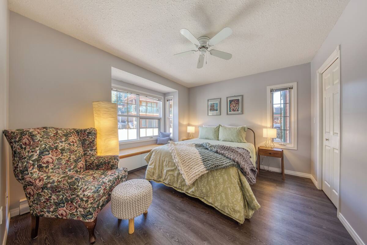 A bedroom with a bed, a floral armchair, a small ottoman, a ceiling fan, and two bedside lamps.