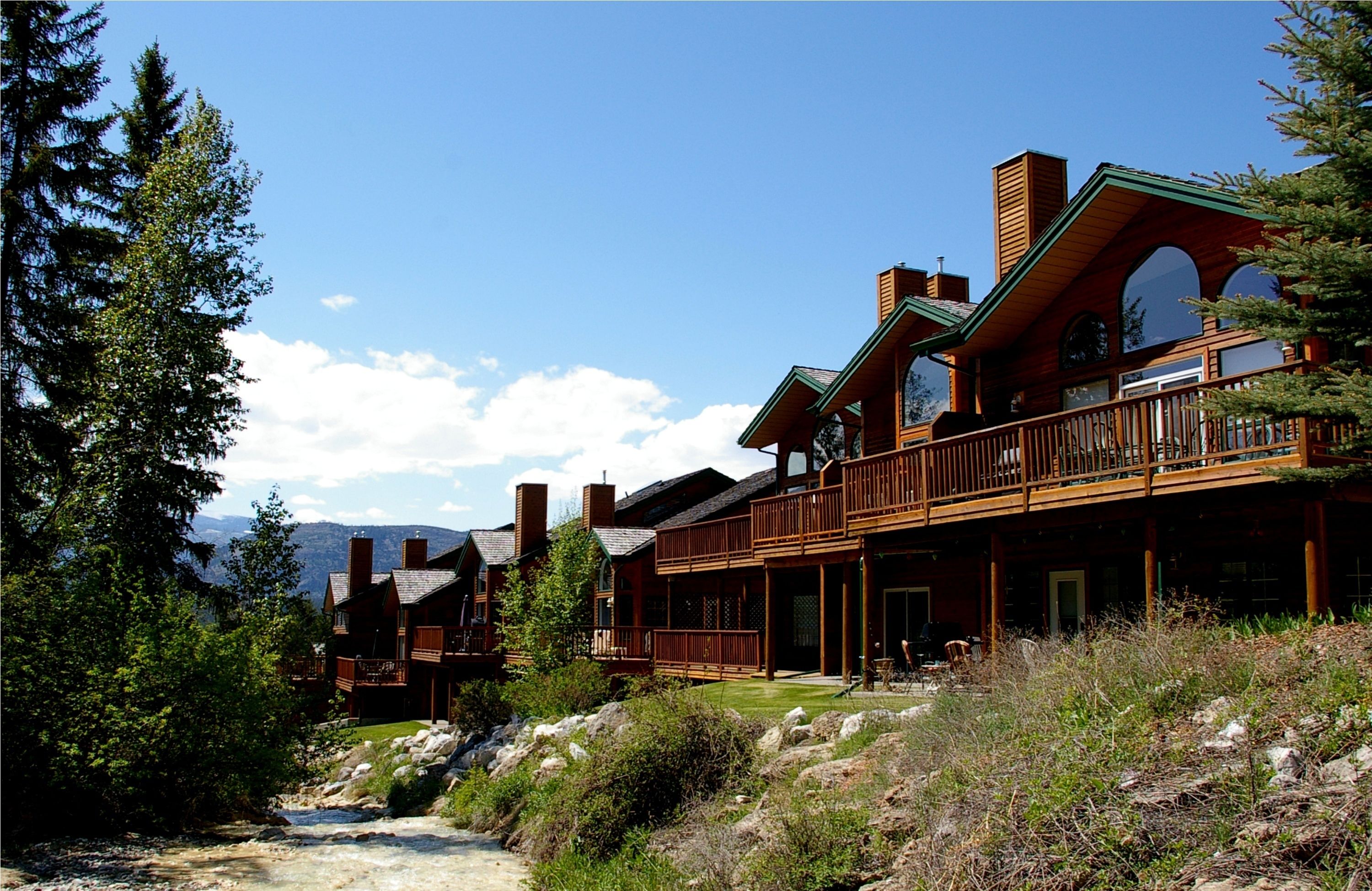 A large wooden lodge with multiple balconies and large windows, surrounded by trees and a rocky landscape.