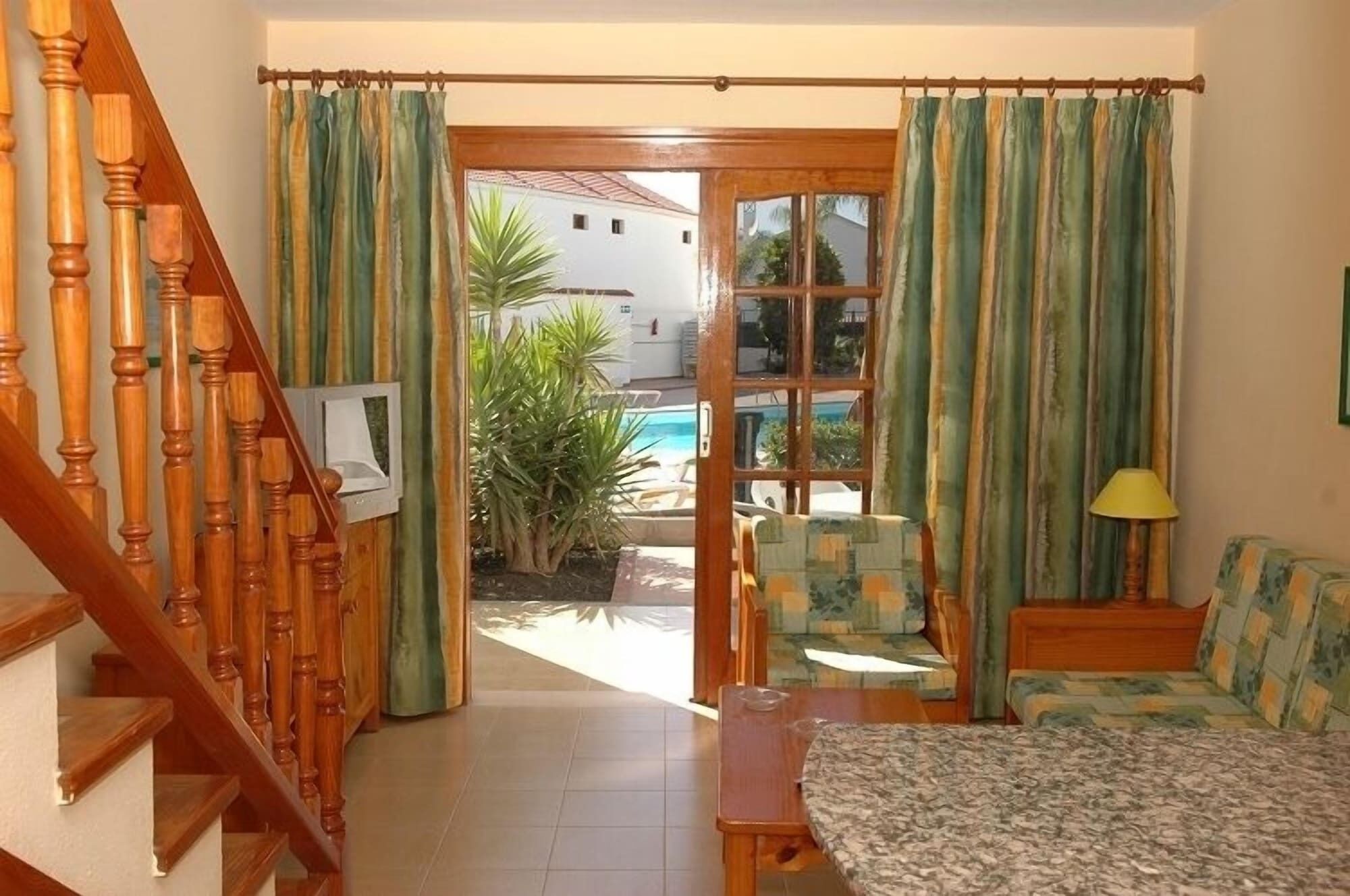 A room with a wooden staircase, a television, a sofa, a table, and a view of an outdoor pool area.