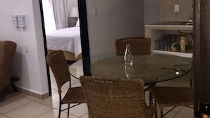 Un'area pranzo con tavolo in vetro e sedie in vimini, una cucina con zanzara piastrellata e un ingresso che porta a una camera da letto con letto e tende.