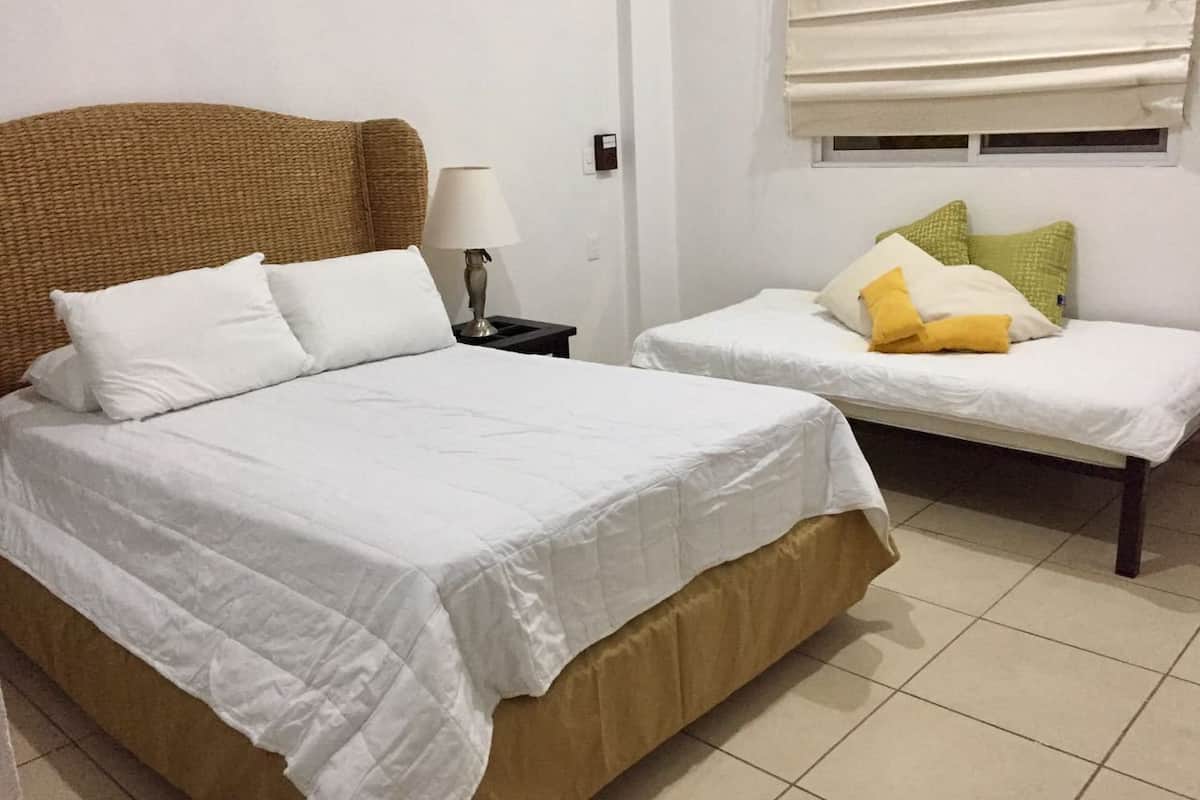 Een hotelkamer met een groot bed, een kleiner bed, een nachtkastje met een lamp en een raam met jaloezieën.