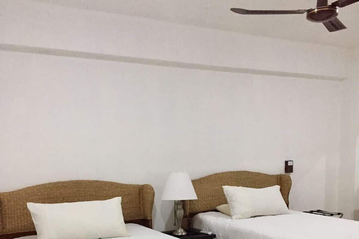 Een hotelkamer met twee bedden, een plafondventilator, een nachtkastje met lamp en een stopcontact.