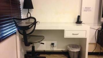 A small, compact room with a white desk, a black office chair, a lamp, a trash bin, and a bed with a blue blanket.