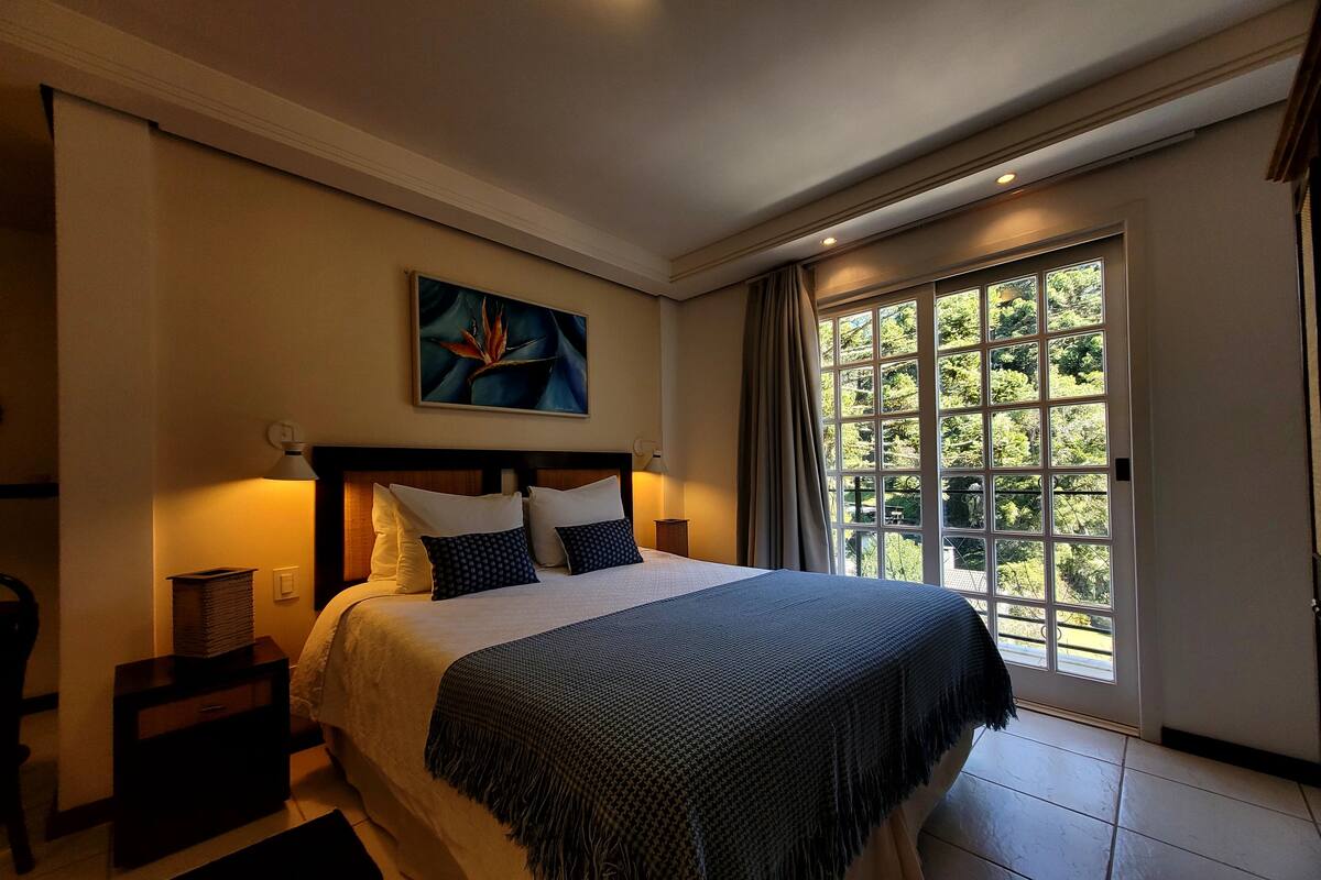 Une chambre avec un grand lit, une table de chevet, une lampe, un tableau au mur et une fenêtre donnant sur des arbres.