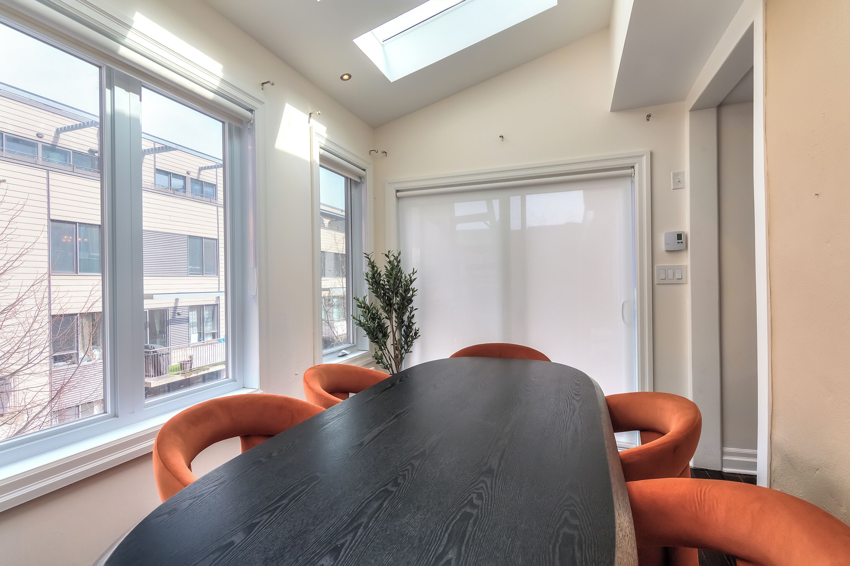 A conference room with a large table, orange chairs, a potted plant, and large windows with a view of buildings outside.