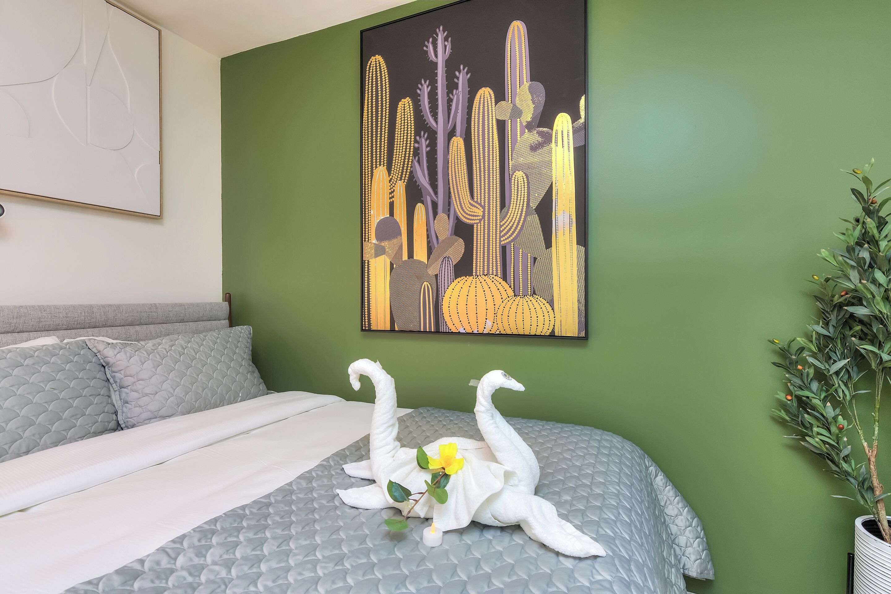 A bed with a gray quilted bedspread, a white pillow, and a decorative swan headboard. A cactus-themed painting is on the wall above the bed.