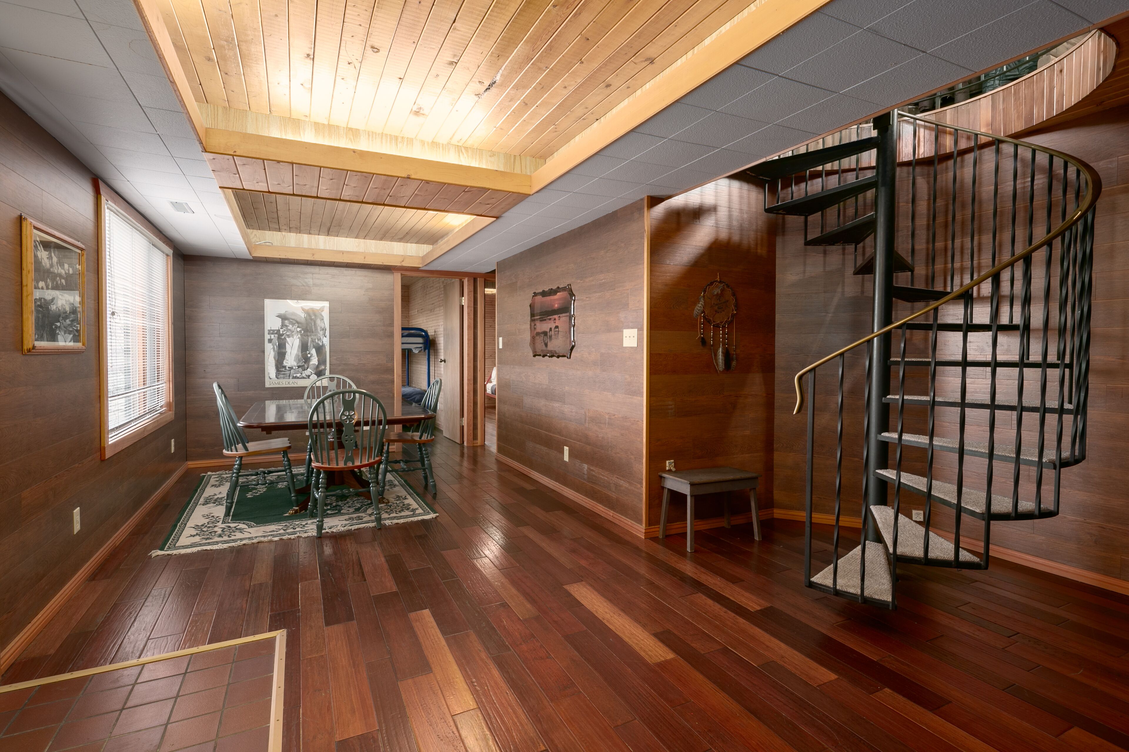 Une pièce spacieuse dotée d’un plancher en bois, d’un coin repas avec des chaises et une table, d’un escalier en colimaçon et d’une grande fenêtre avec des stores.