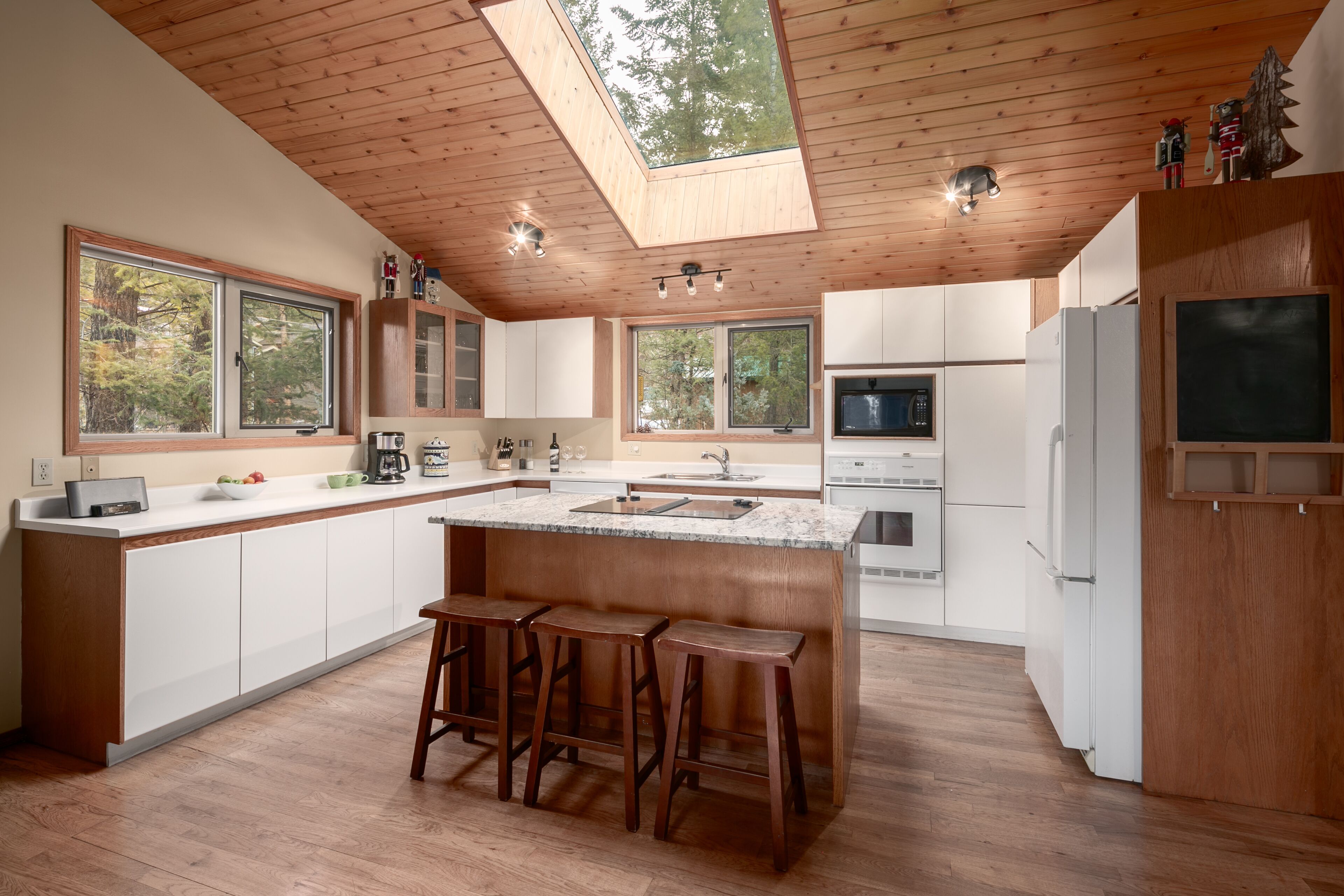 A modern kitchen with a central island, wooden ceiling, large windows, and a skylight.