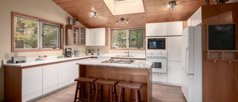 A modern kitchen with a central island, wooden ceiling, large windows, and a skylight.