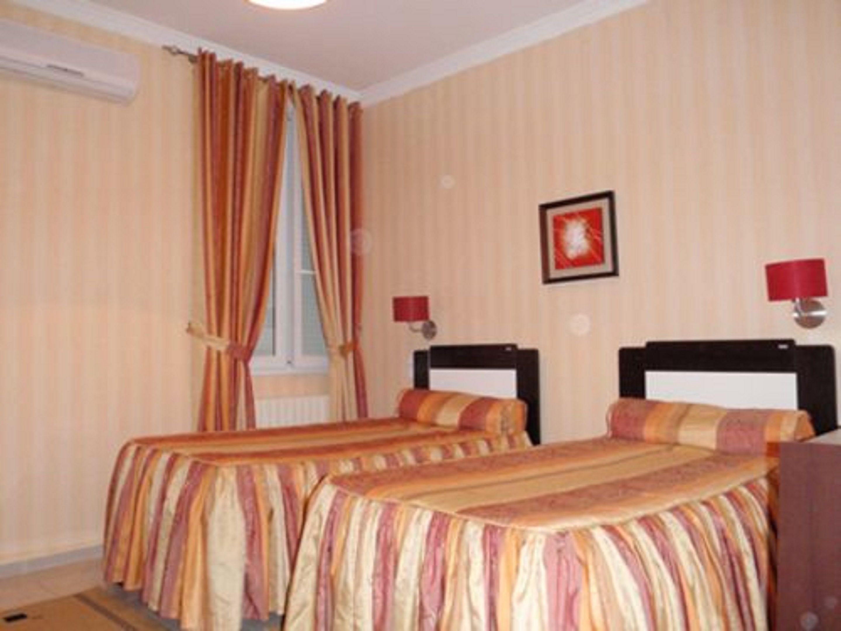 Een hotelkamer met twee bedden, gestreept beddengoed, een raam met gordijnen, een wandlamp en een inlijsting foto.