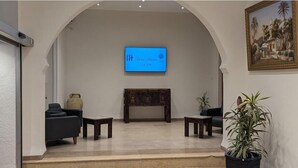 A lobby with a blue screen displaying text, a traditional wooden cabinet, two armchairs, a small table, and a potted plant. -  Hôtel Audin (Algiers)