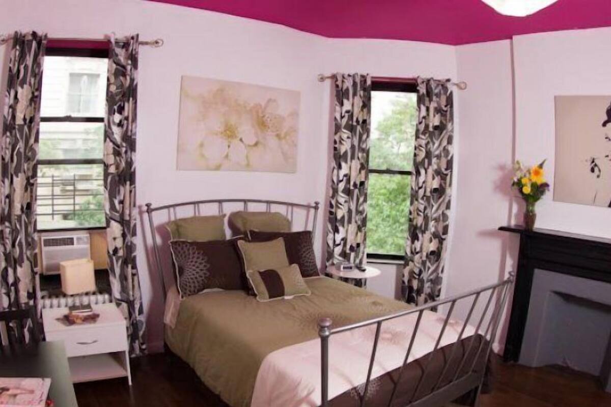 Quarto com estrutura de cama de metal, mesa de cabeceira, cadeira e janela com cortinas estampadas.