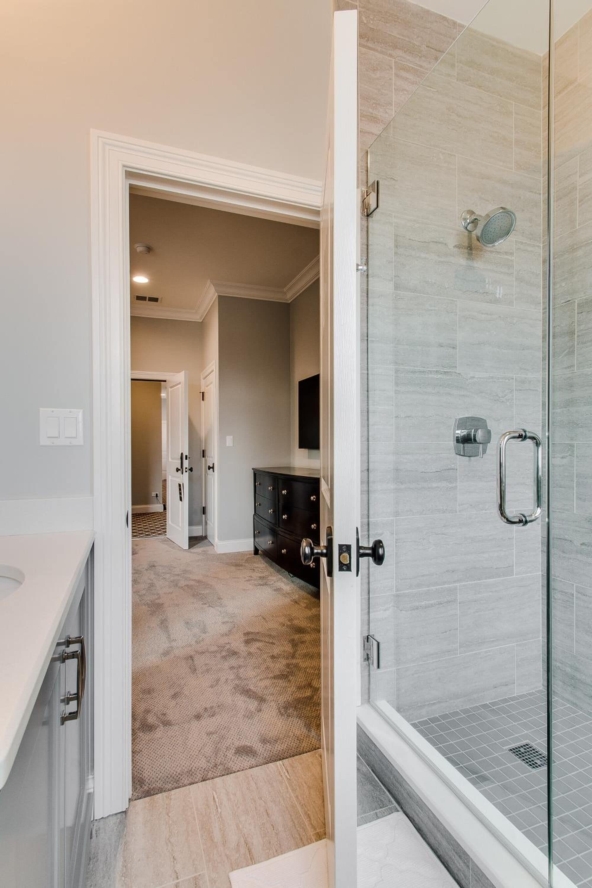A modern bathroom with a glass shower enclosure, a white vanity, and a beige carpeted hallway leading to a bedroom.