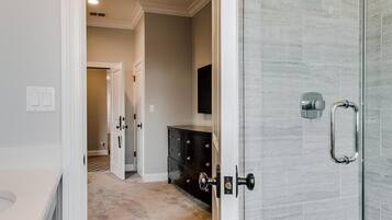 A modern bathroom with a glass shower enclosure, a white vanity, and a beige carpeted hallway leading to a bedroom.