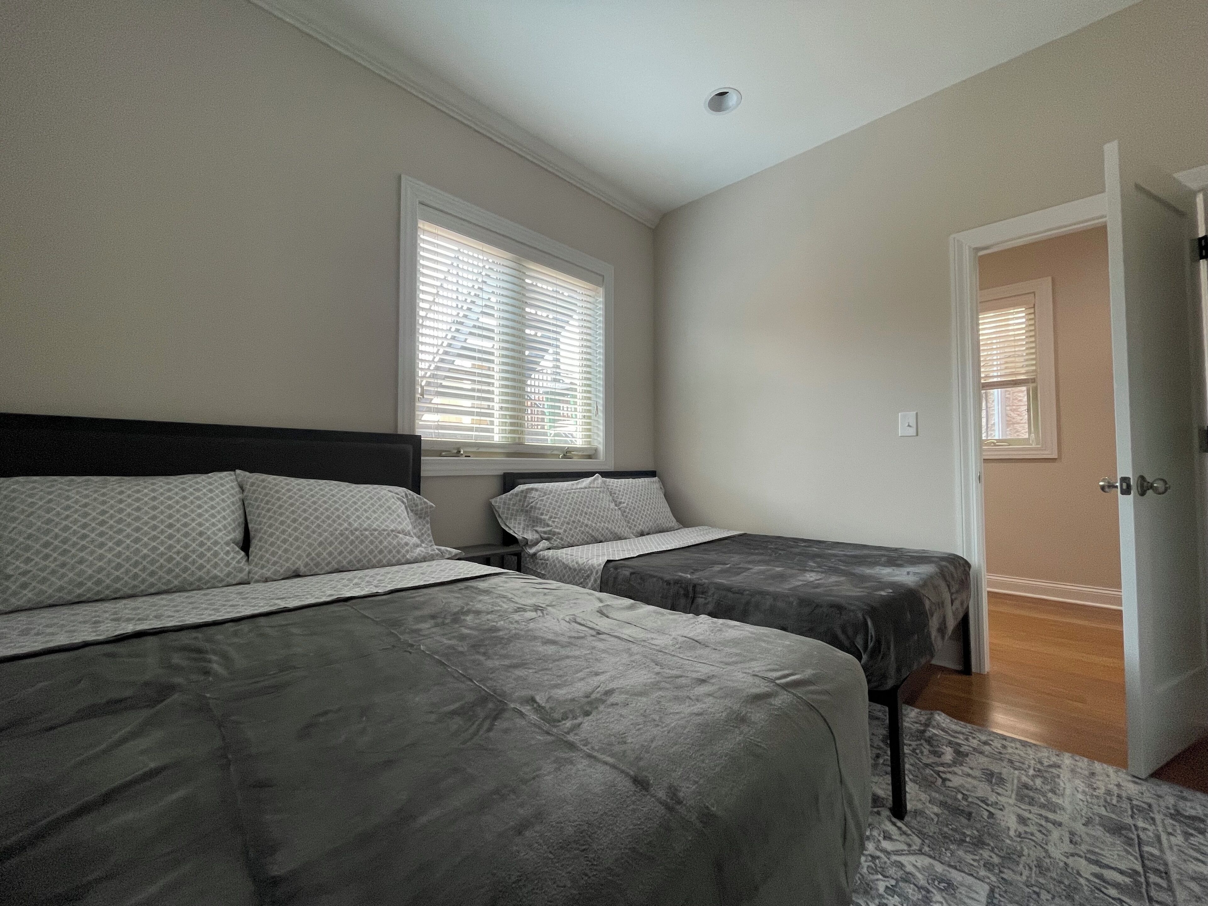 A bedroom with two beds, a window with blinds, a door, and a ceiling light.