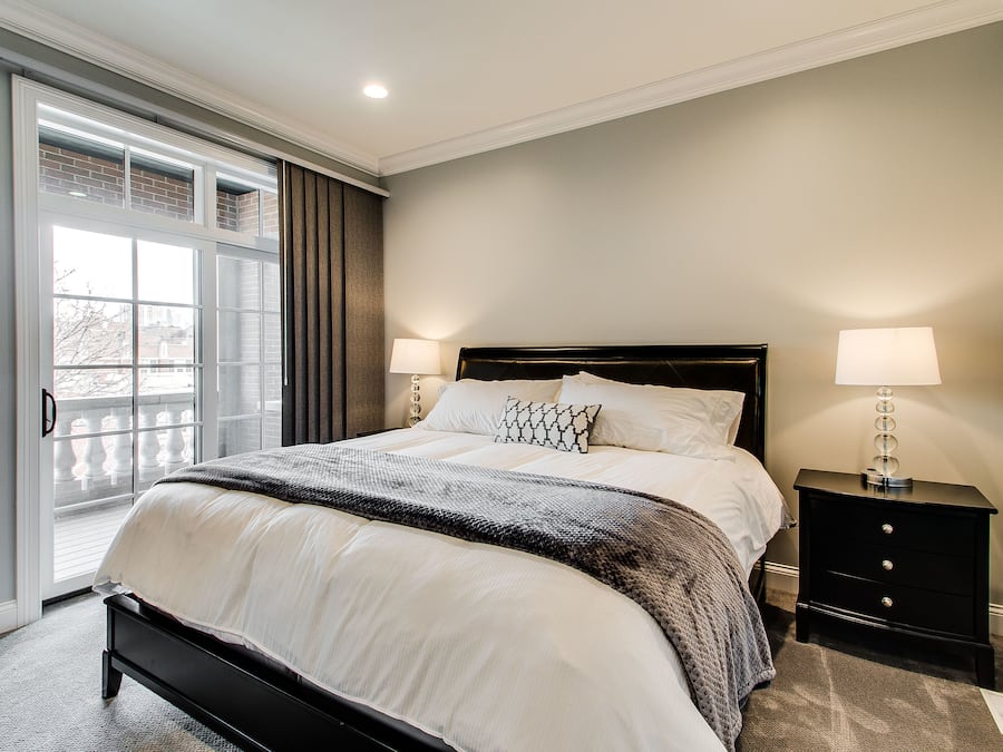A bedroom with a large bed, two bedside tables with lamps, a window with blinds, and a view of the outside.