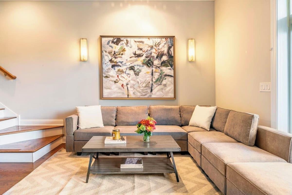 Una sala de estar con un sofá seccional, una mesa de centro con libros y un jarrón de flores, y un cuadro abstracto grande en la pared.