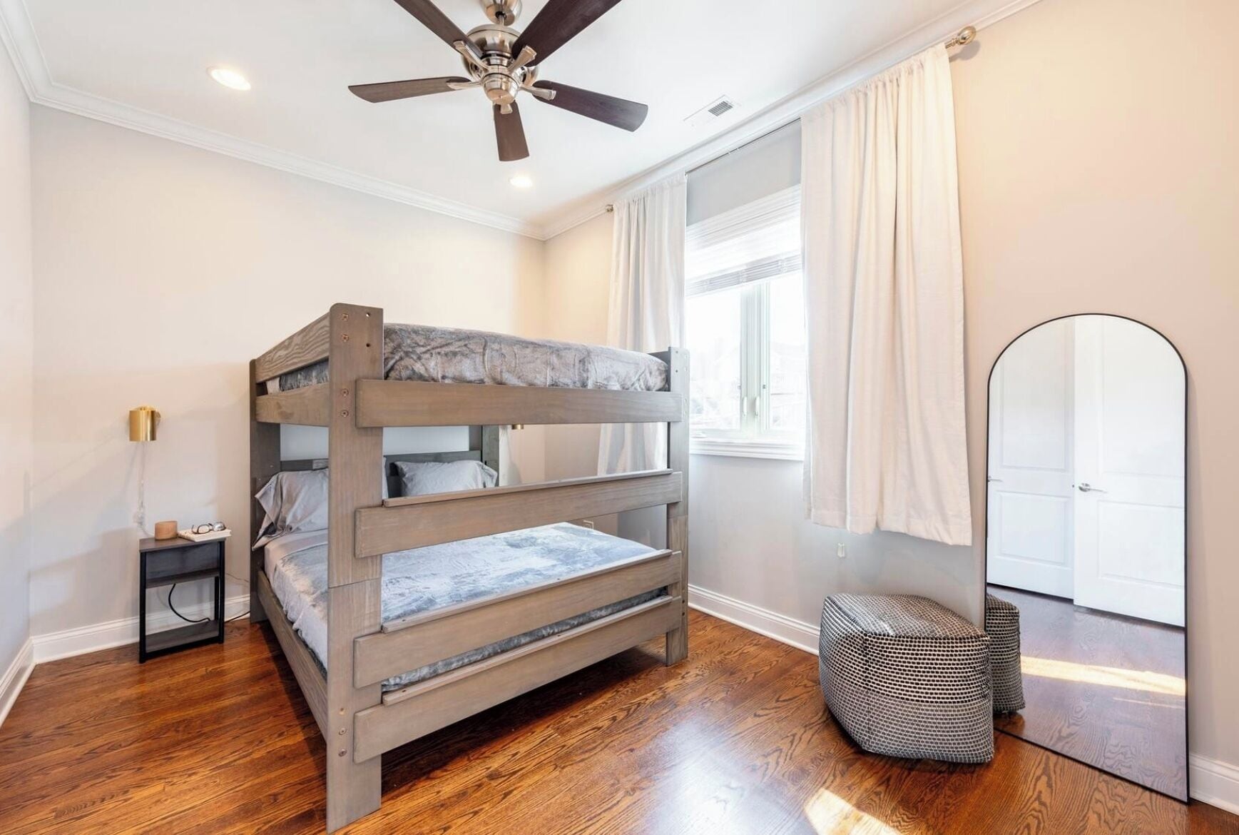 A bunk bed room with a ceiling fan, a bedside table with a lamp, a window with curtains, and a large mirror.
