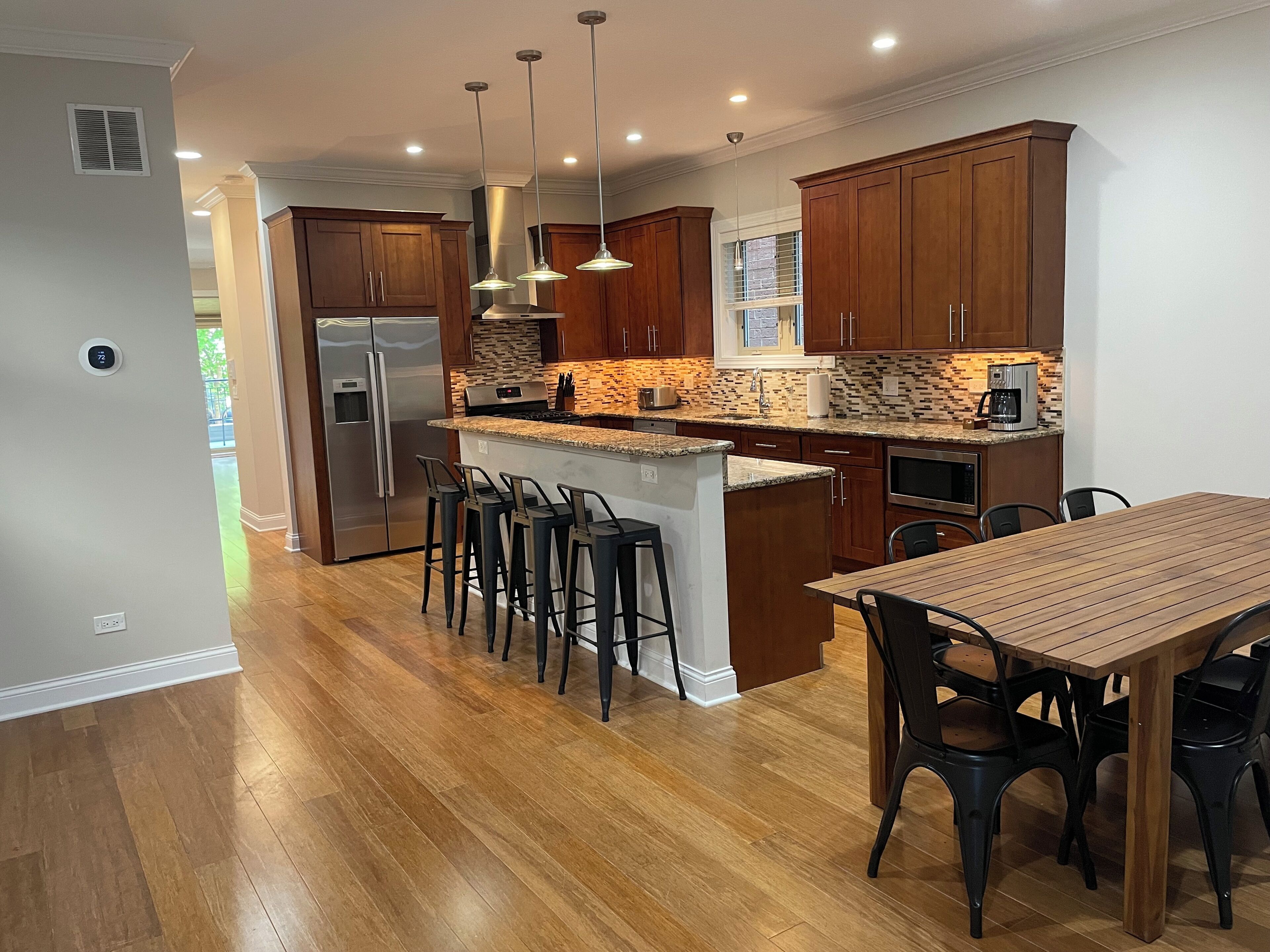 A modern kitchen with wooden cabinets, a stainless steel refrigerator, a built-in island with bar stools, and a dining table with chairs.