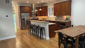 A modern kitchen with wooden cabinets, a stainless steel refrigerator, a built-in island with bar stools, and a dining table with chairs.
