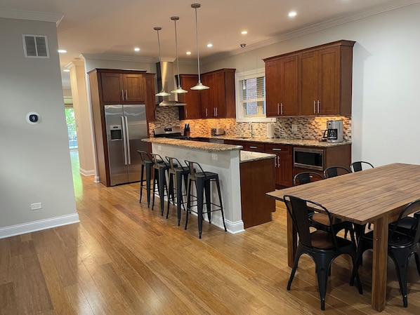 A modern kitchen with wooden cabinets, a stainless steel refrigerator, a built-in island with bar stools, and a dining table with chairs.