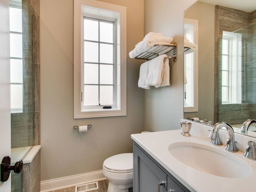 A bathroom with a white sink, two faucets, a mirror, and a window above the toilet.