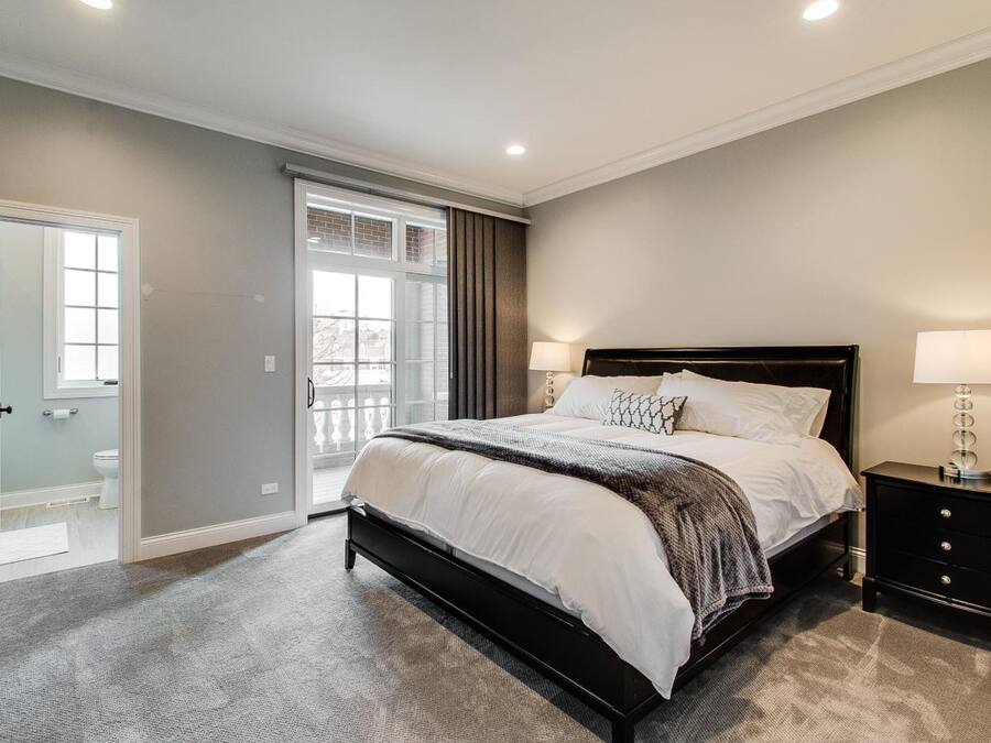 A bedroom with a large bed, a nightstand, a window with curtains, and a door leading to another room.