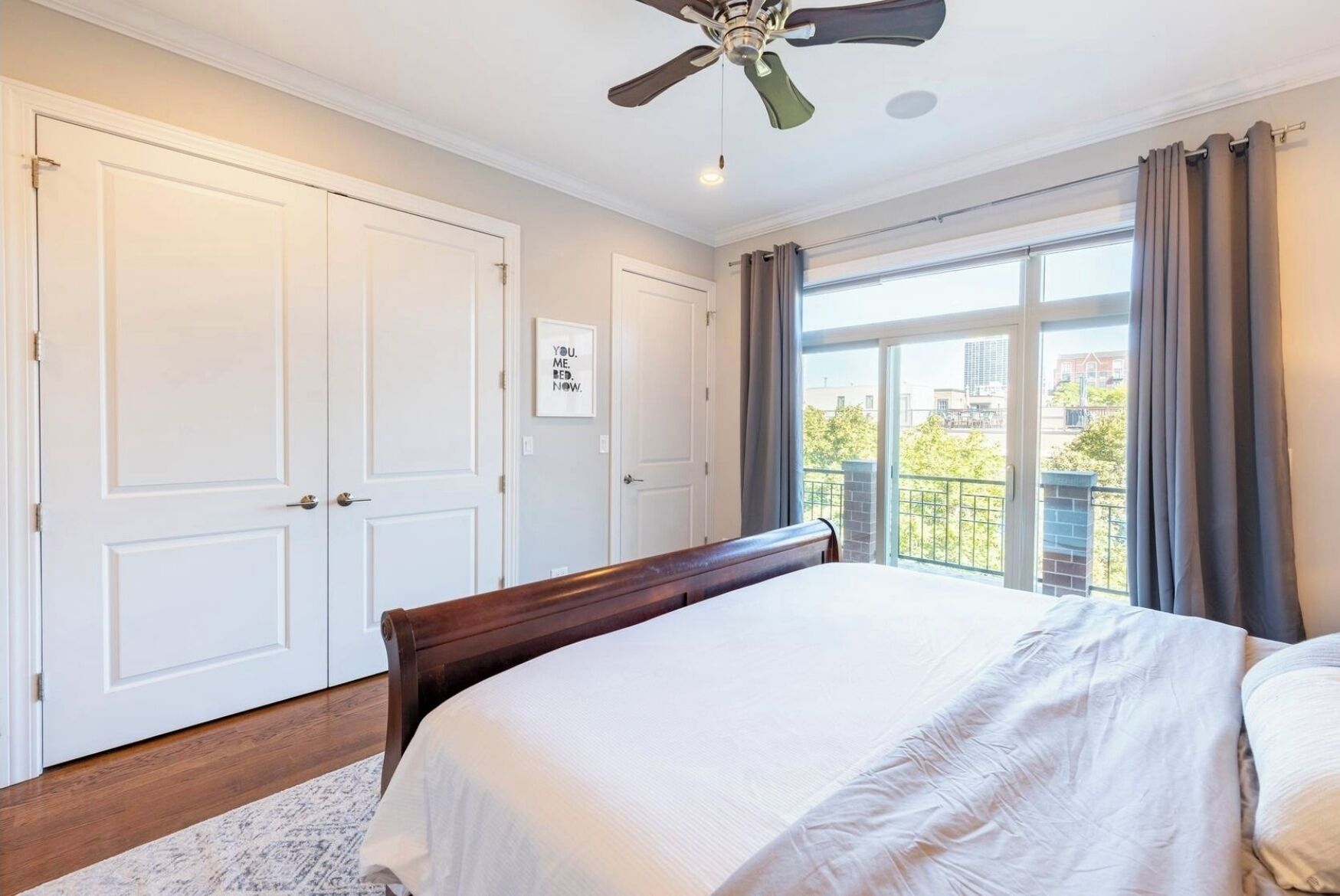 A bedroom with a large bed, a ceiling fan, white doors, and a view of the outside through a window.