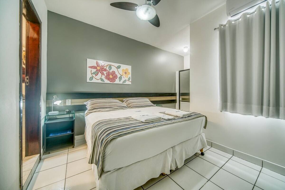 A modern hotel room with a bed, bedside tables, a ceiling fan, a wall-mounted air conditioner, and a window with curtains.