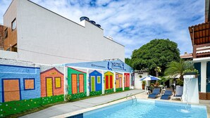 Una piscina con edificios de colores al fondo, cielo despejado y un árbol.