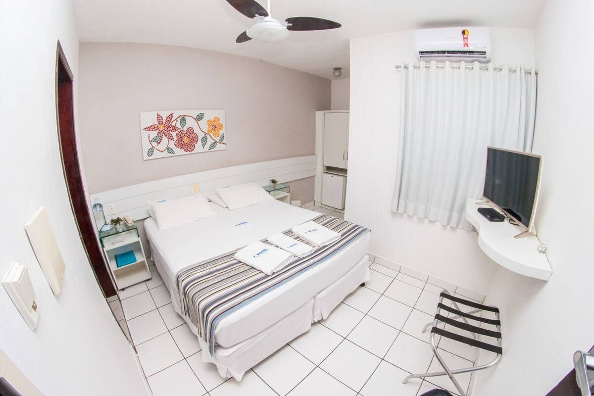 A hotel room with a bed, a desk with a computer, a ceiling fan, an air conditioner, and a window with curtains.