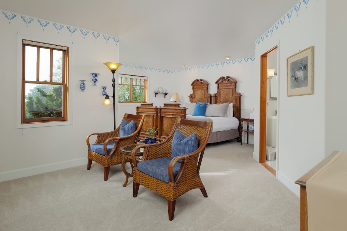 A room with a bed, two wicker chairs with cushions, a lamp, and a wooden cabinet.