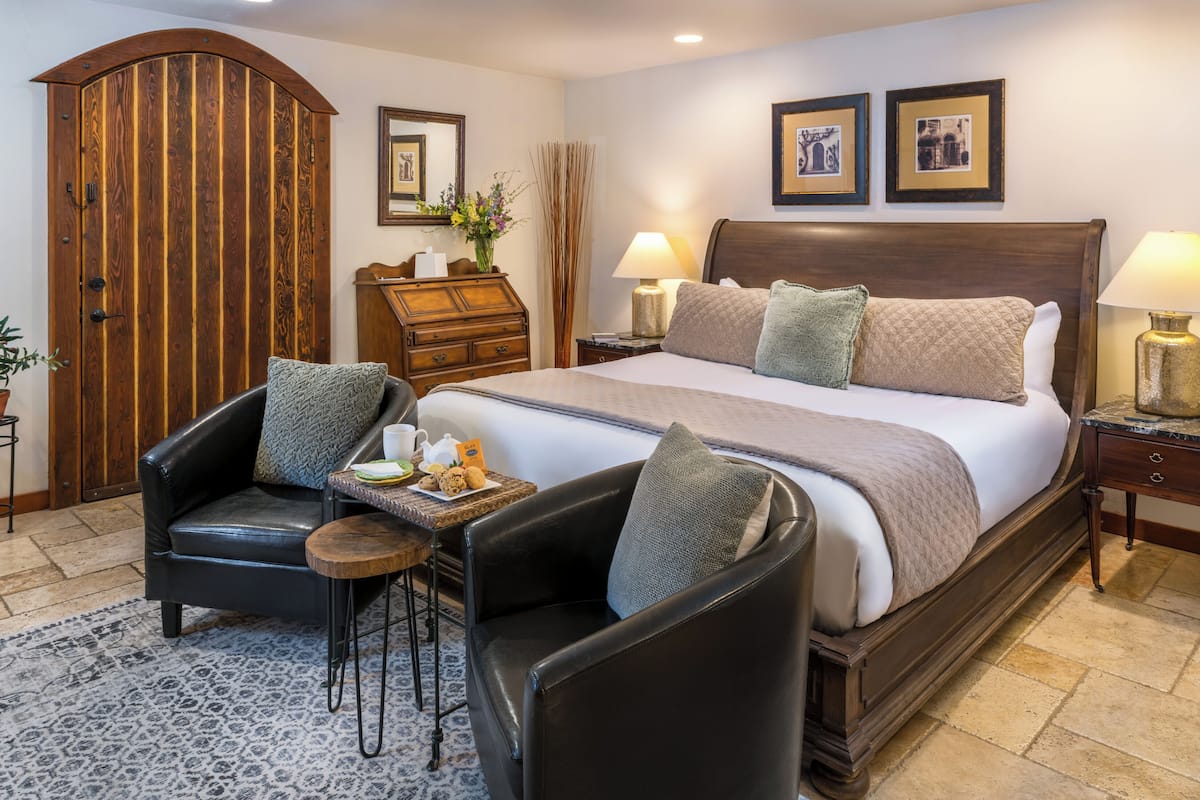 A hotel room with a large bed, two side tables with lamps, a wooden wardrobe, a black leather armchair, and a small table with a tray of breakfast items.