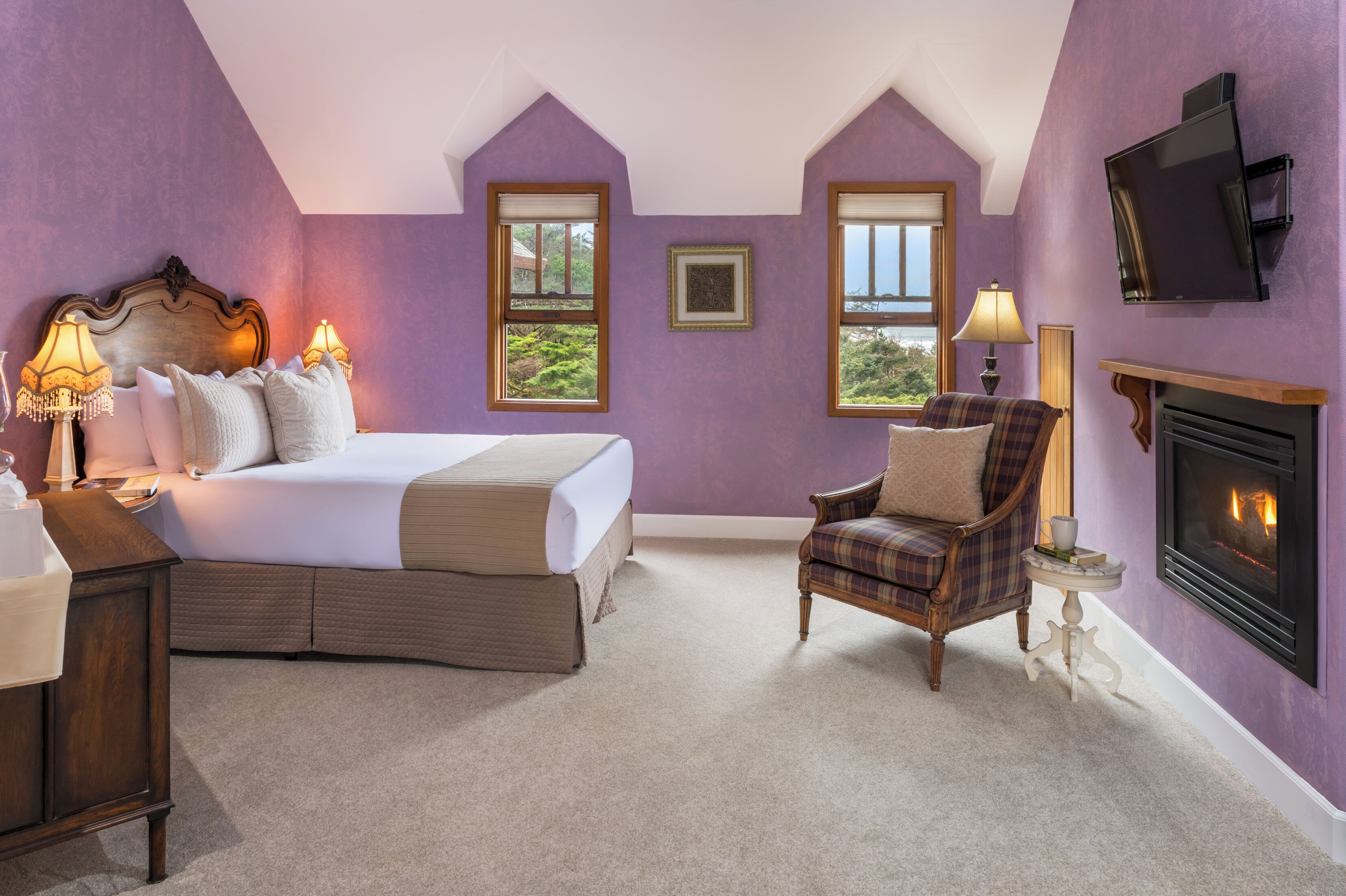 A bedroom with a bed, a chair, a fireplace, and two windows.