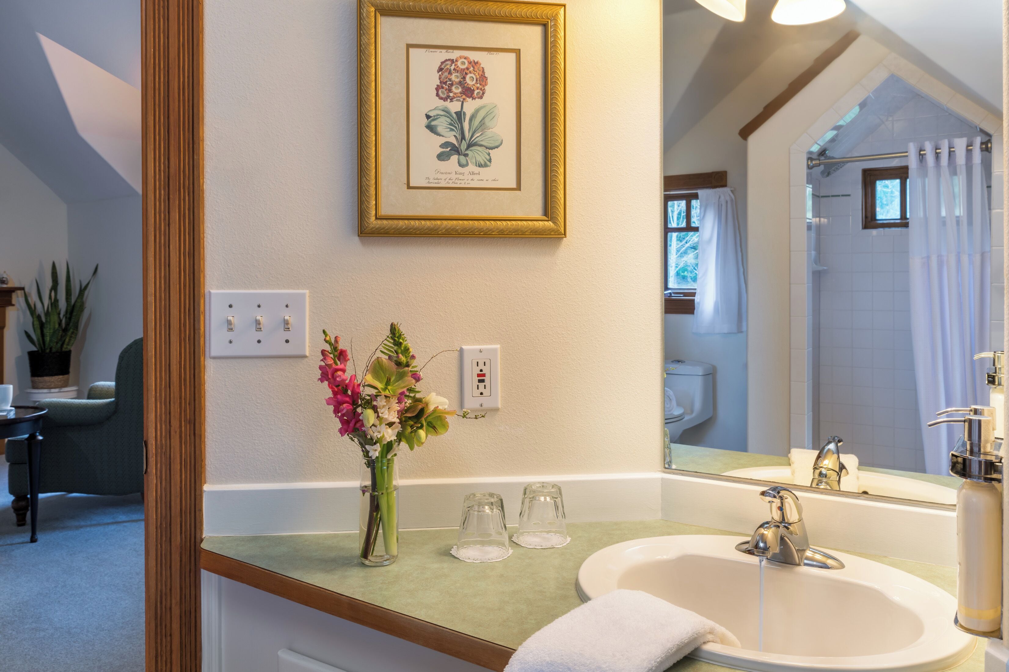 Une salle de bain avec un lavabo, un miroir et une douche. Un tableau botanique encadré orne le mur, et un vase rempli de fleurs est posé sur le comptoir.