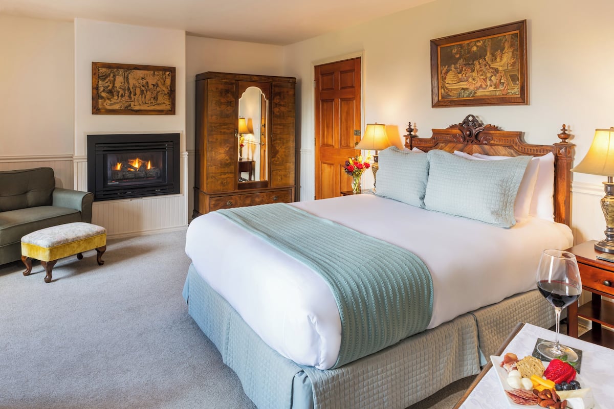 A hotel room with a large bed, a fireplace, a wooden wardrobe, a nightstand with a lamp, and a dining table with a wine glass and snacks.