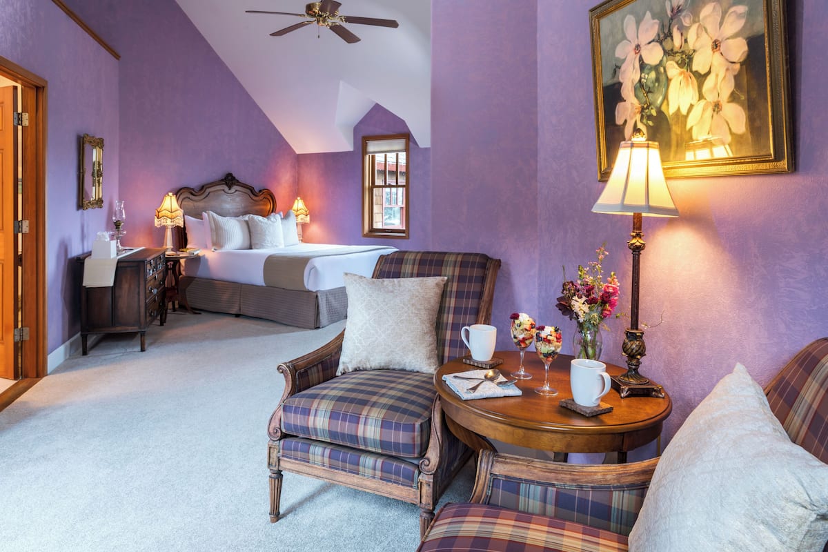 A bedroom with a bed, a chair, a table with a lamp, and a painting on the wall.