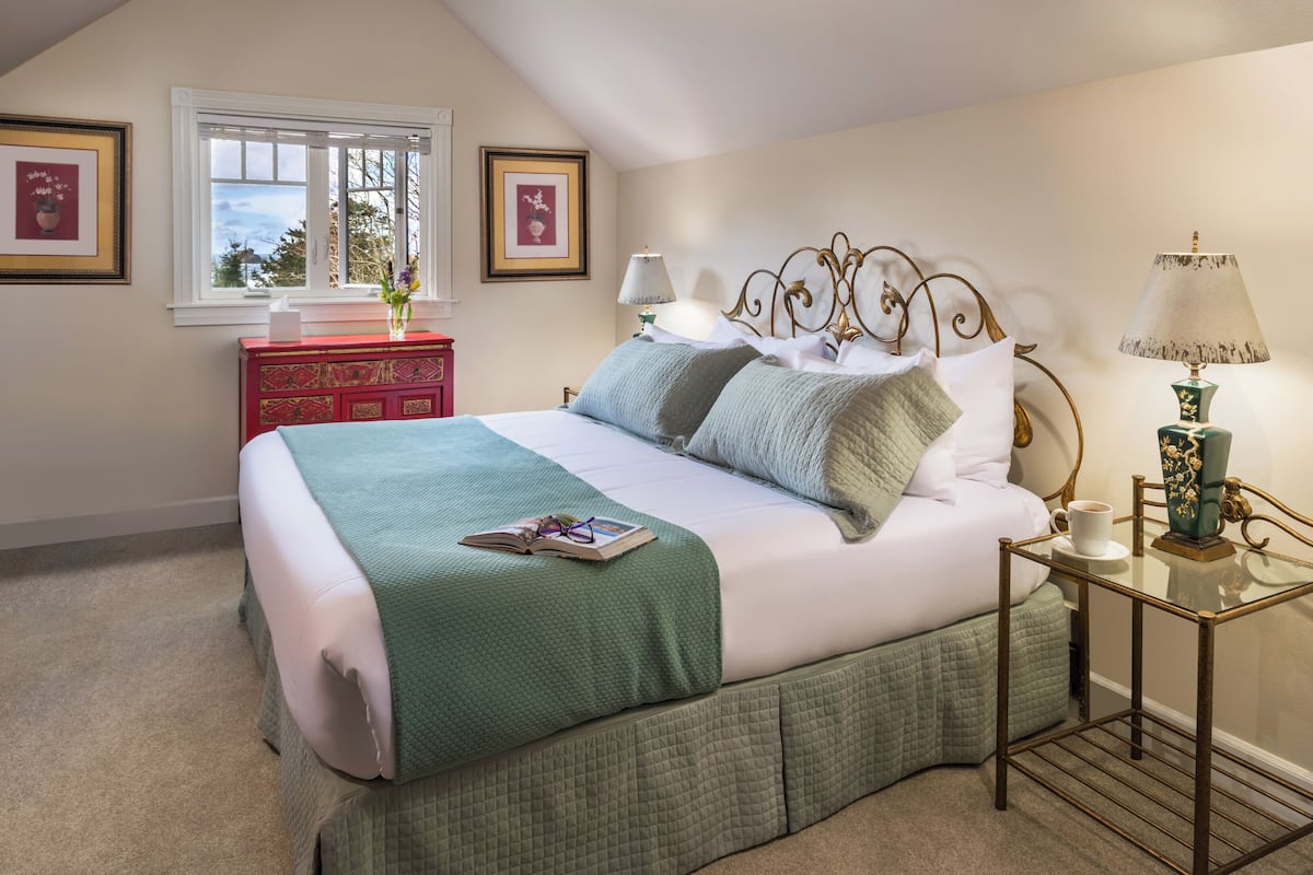 A neatly made bed with a green bedspread and pillows, a bedside table with a lamp, and a dresser with a vase of flowers.