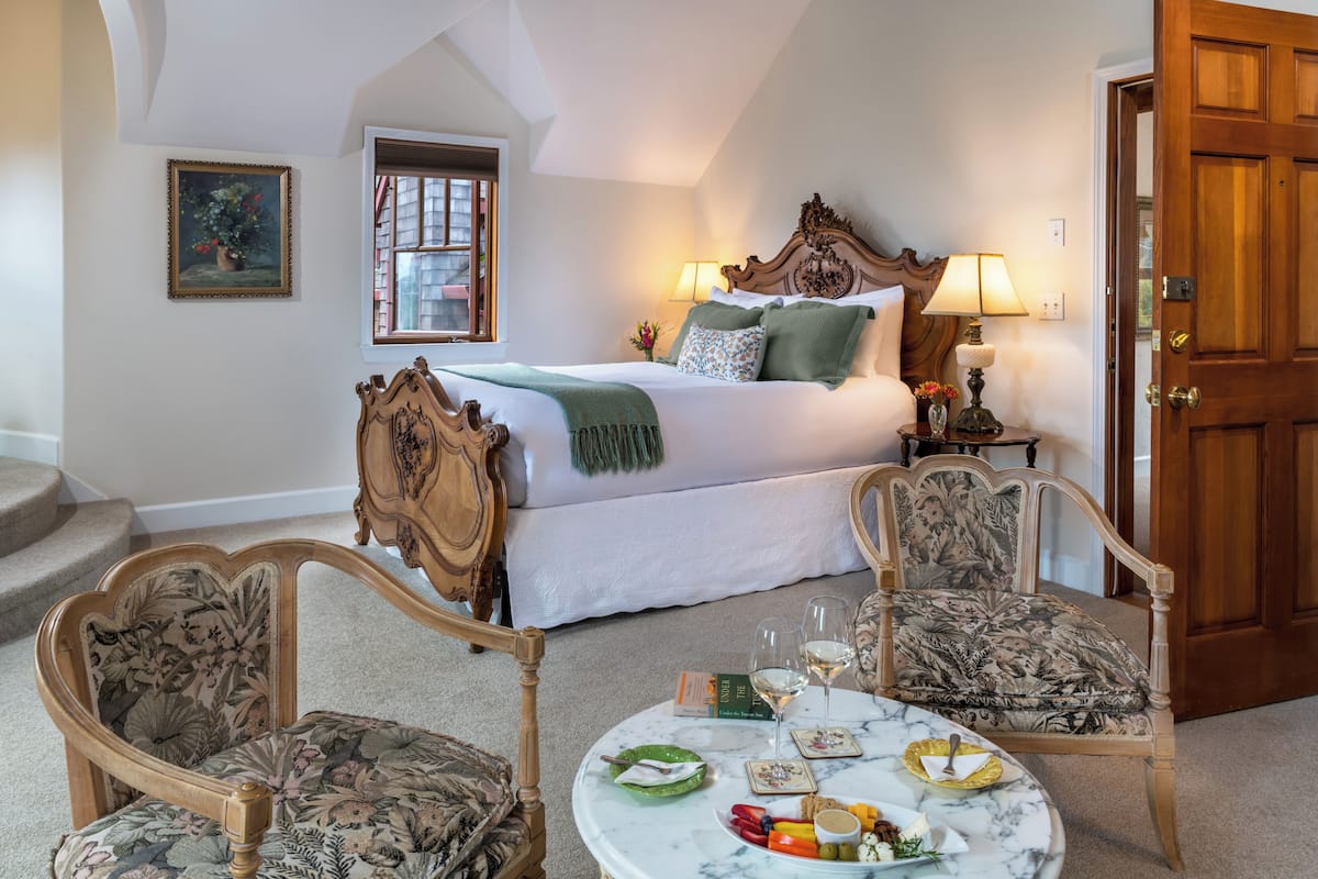 A bedroom with a large bed, two chairs, a bedside table with a lamp, a window, and a door.
