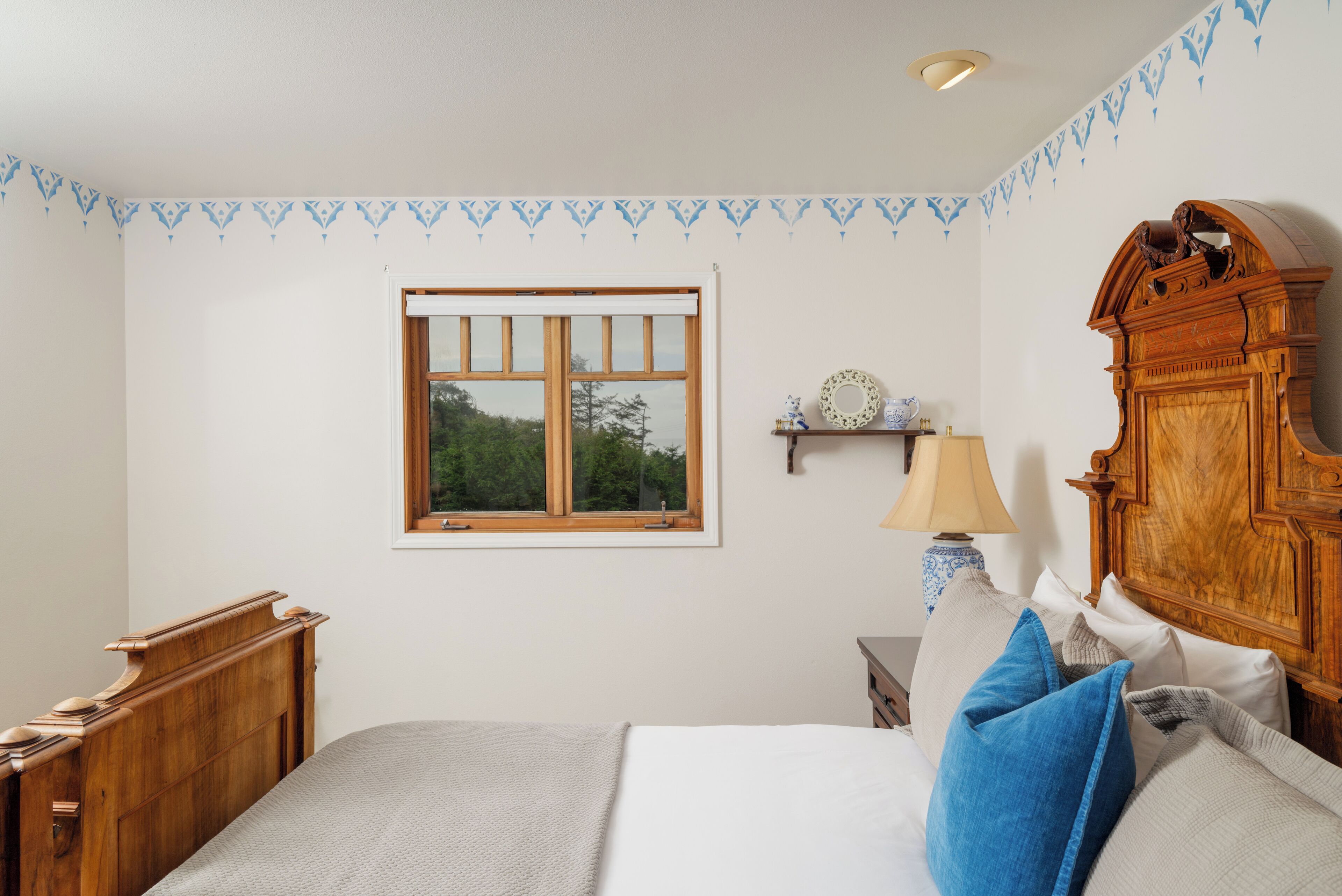 A bedroom with a wooden headboard, a bed with multiple pillows, a nightstand with a lamp, a window with a view of trees, and a decorative wall border.