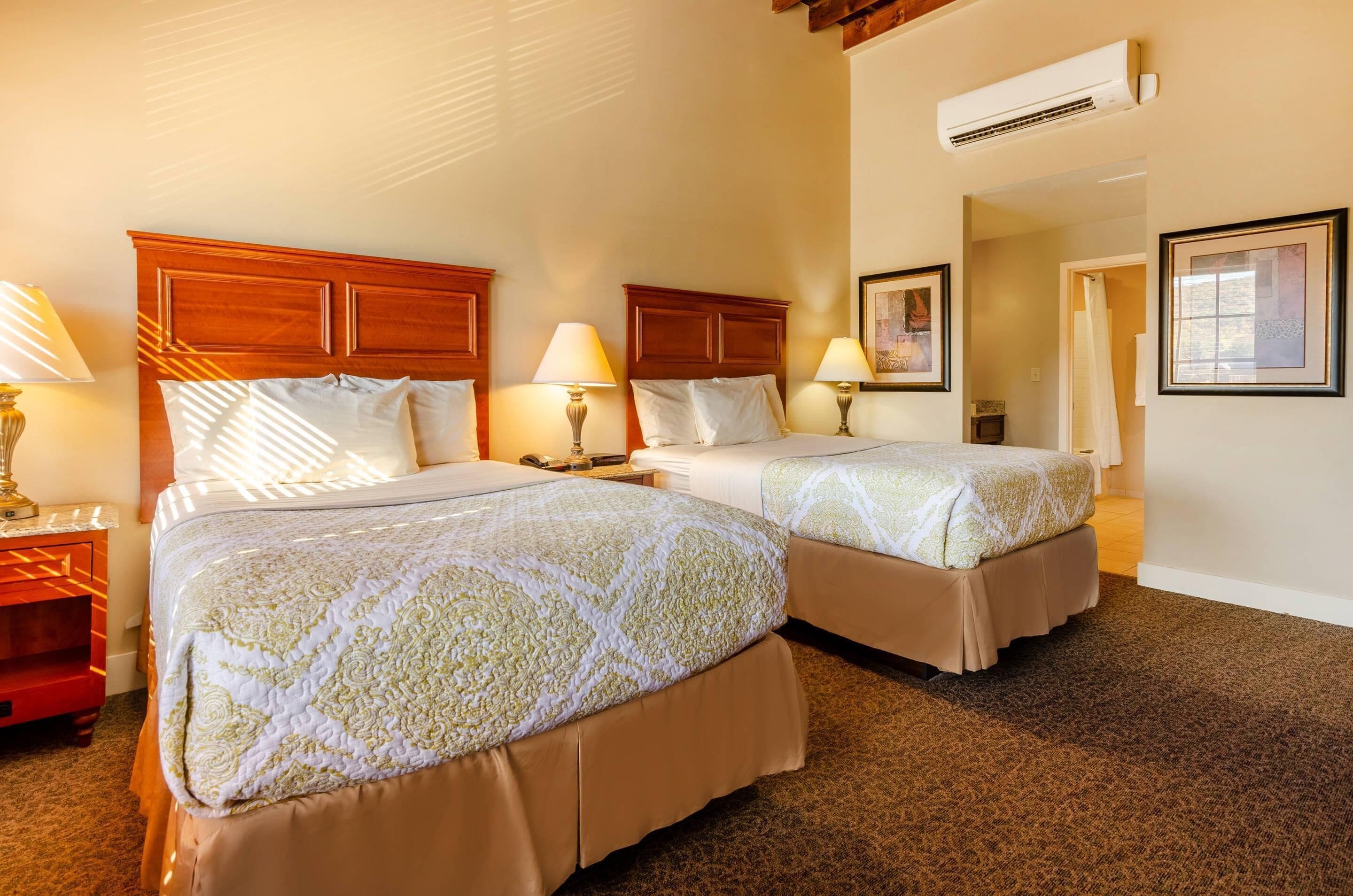 A hotel room with two beds, wooden headboards, bedside lamps, a carpeted floor, and an air conditioning unit.