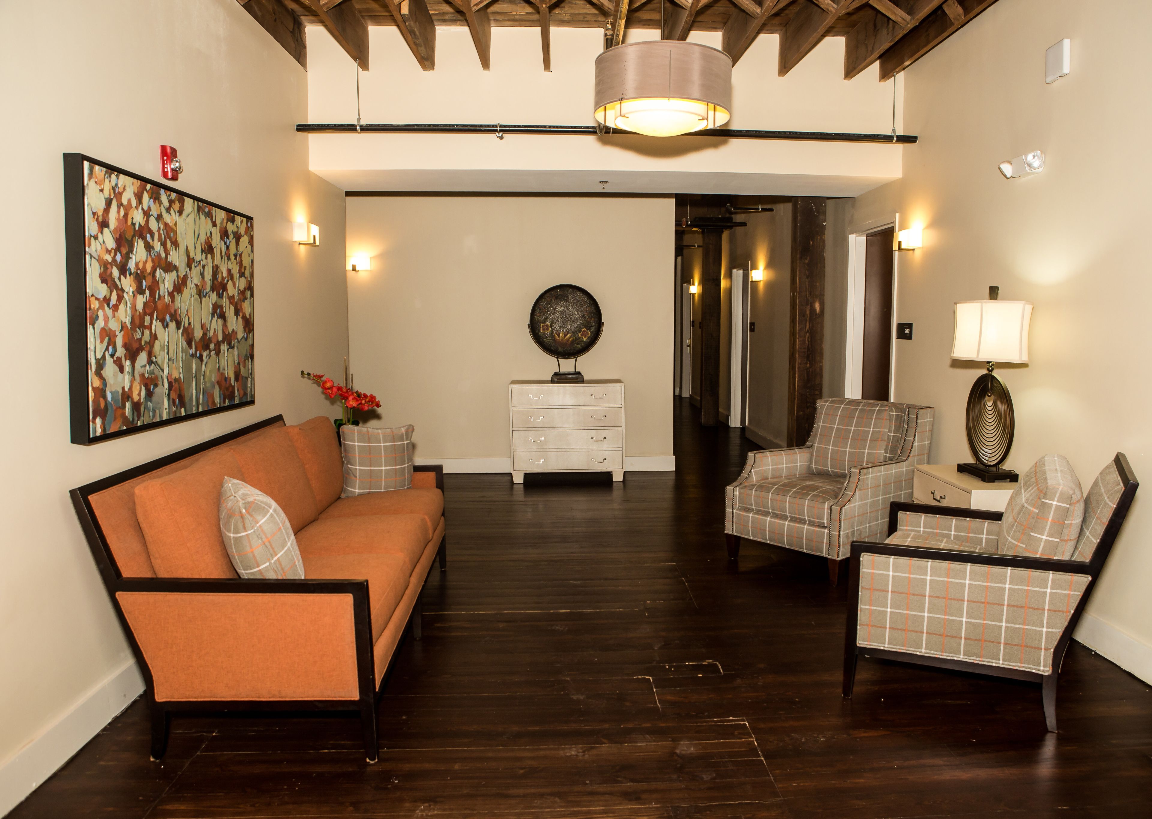 Un salon comprenant un canapé, deux fauteuils, une table d’appoint, une lampe et une assiette décorative. Un tableau est accroché au mur, et un couloir mène à une autre pièce.