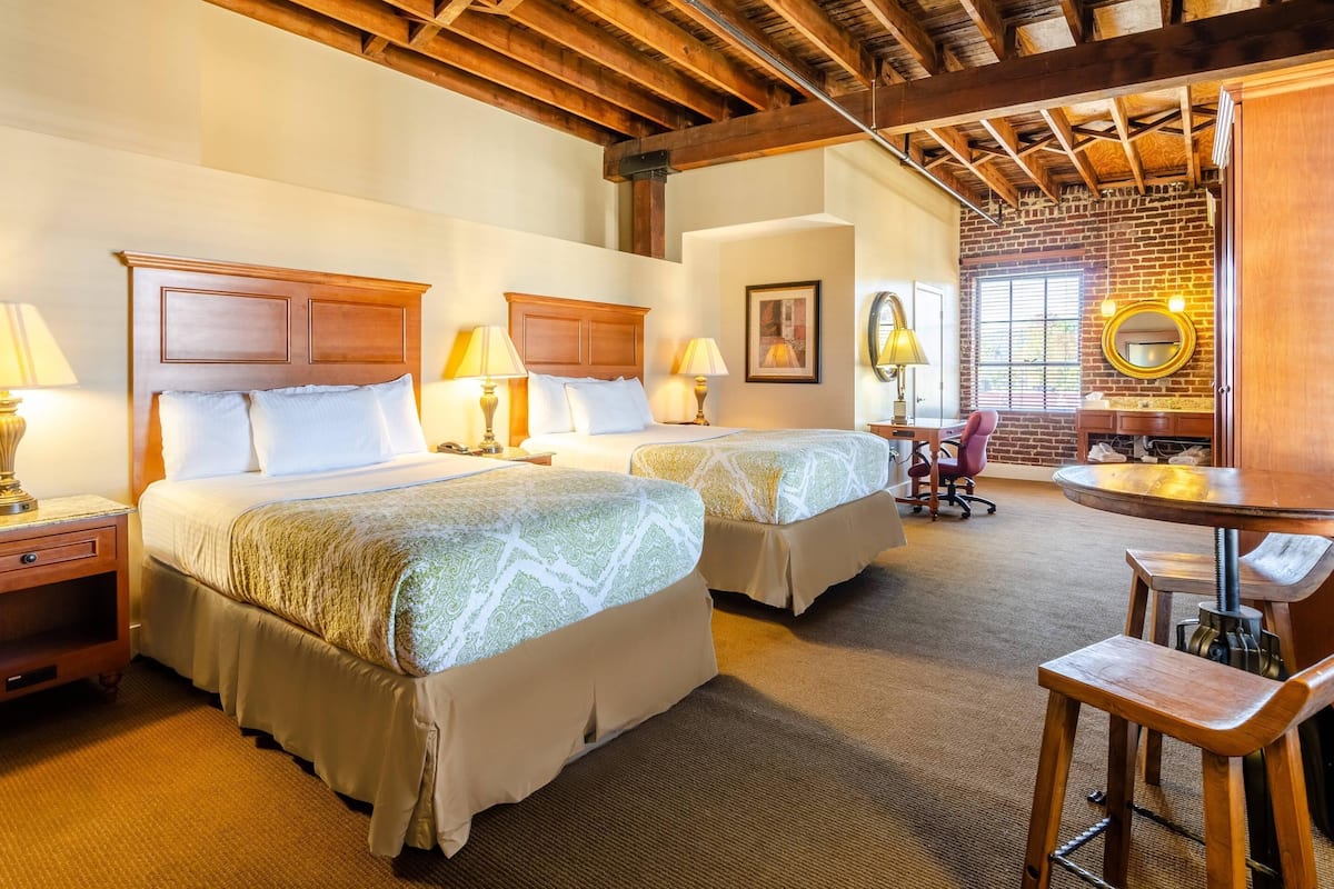 A hotel room with two beds, wooden headboards, and a brick wall. There is a round table with a mirror and a chair.