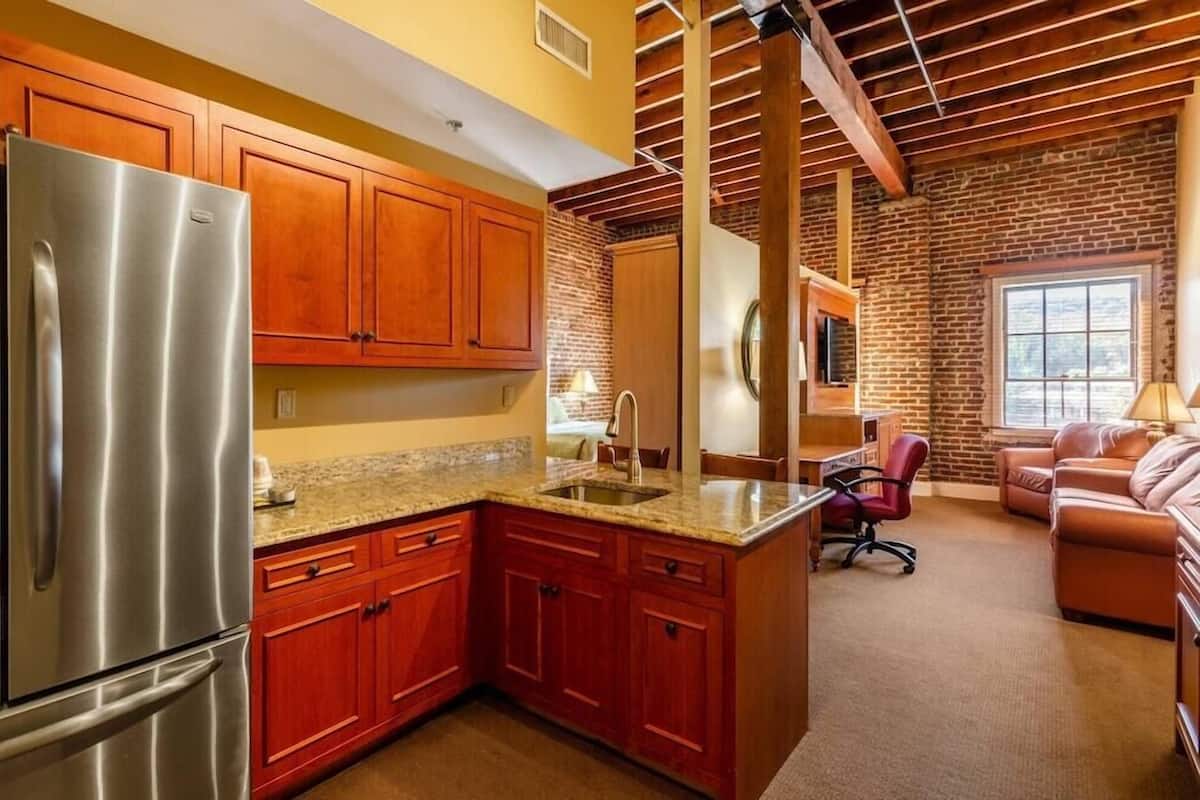 A kitchen with a stainless steel refrigerator, wooden cabinets, granite countertops, and a brick wall.
